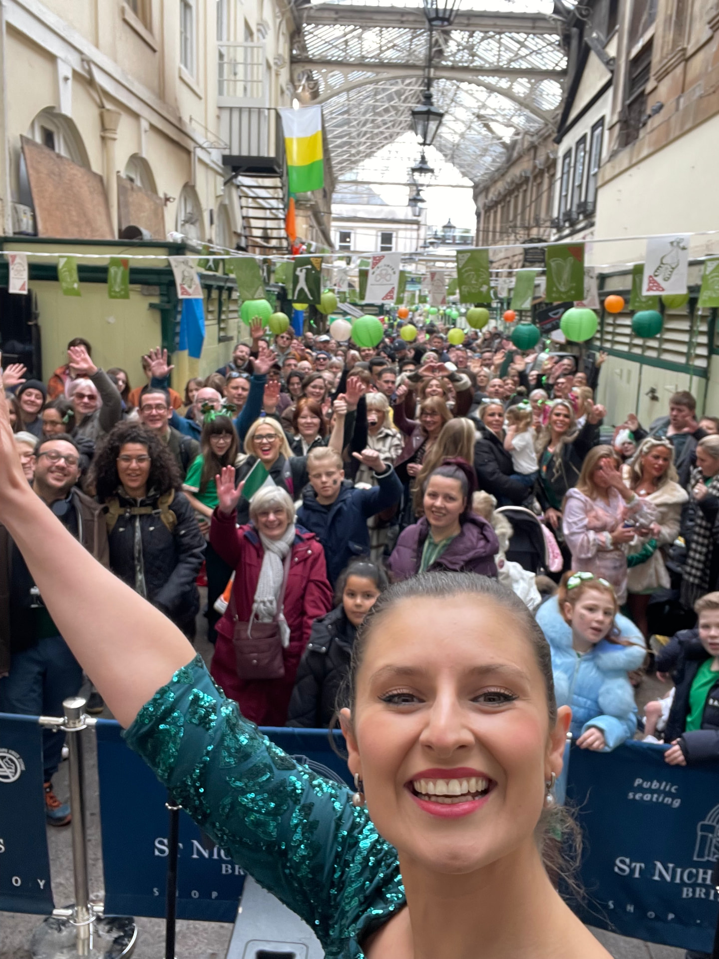 Shannon Kitchen in an emerald green sequined dress in a market arcade decorated with Irish bunting, one arm raised towards a cheering crowd behind her