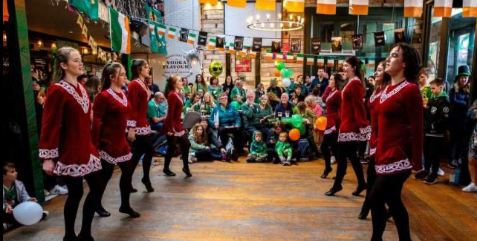 An Irish dance troupe in maroon velvet dance dresses with white Celtic embroidery performing on a wooden floor, watched by an audience in a hall decorated with Irish tricolour flags and bunting