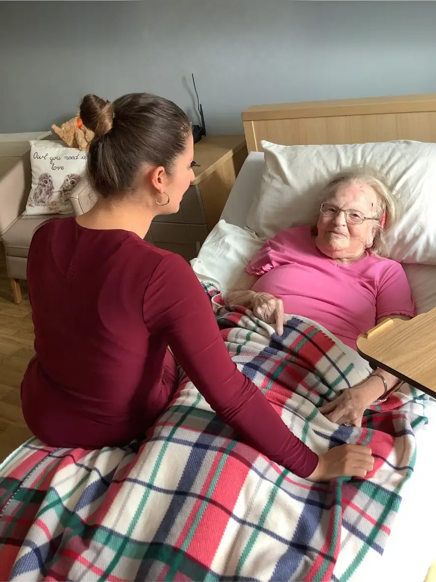 Shannon Kitchen seated on a chair beside a care bed, talking with an elderly resident