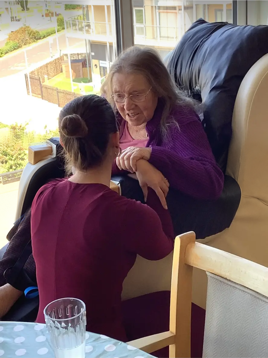 Shannon Kitchen leaning in and holding the hands of an elderly resident seated in an armchair by a window