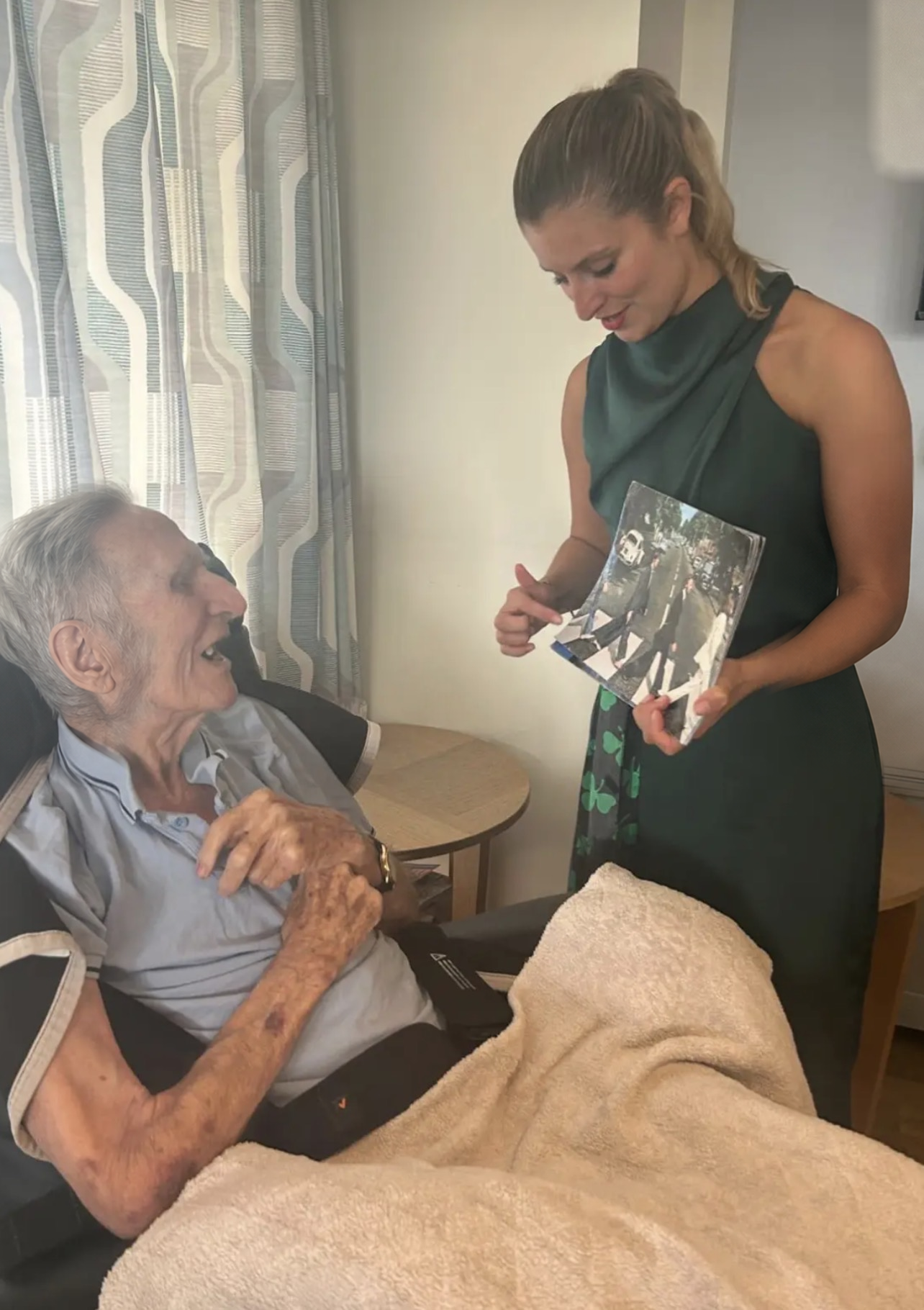 Shannon Kitchen standing and showing an Abbey Road photo album to an elderly resident seated in an armchair